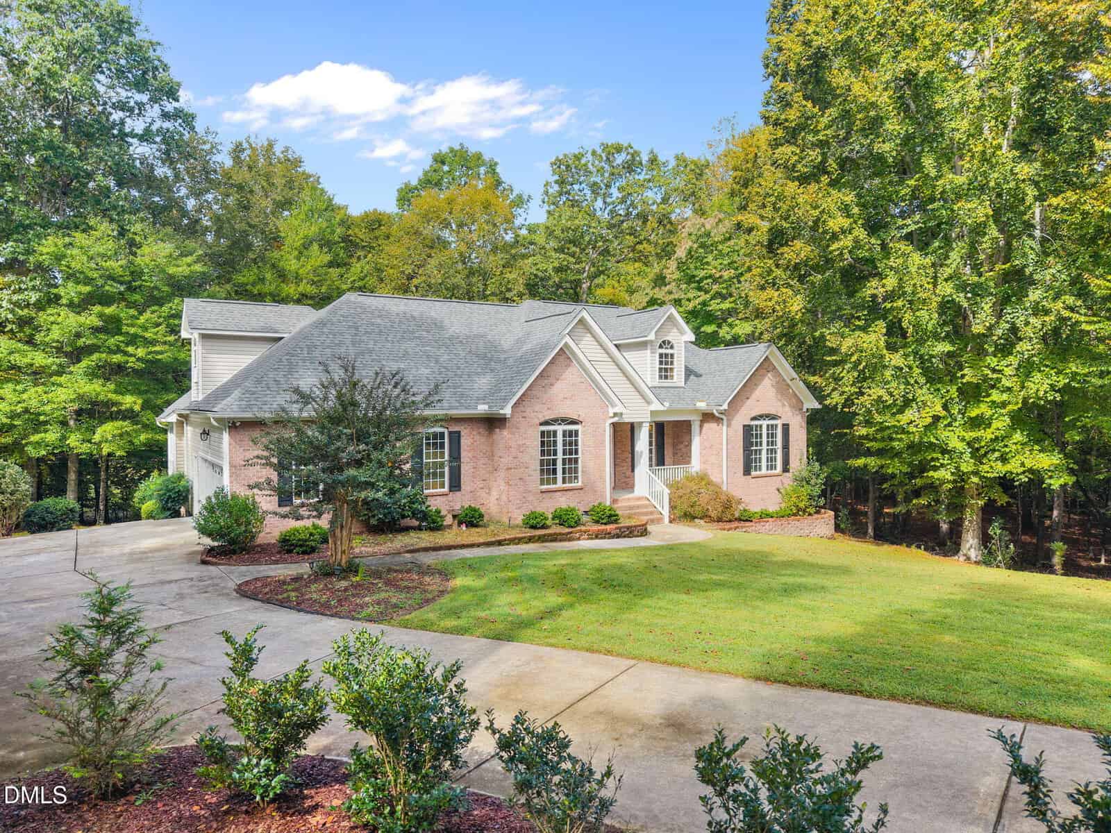 A one-story brick house with gray roofing sits on a well-maintained lawn surrounded by trees. A curved driveway leads to the front steps, and shrubs are planted along the walkway and near the house.