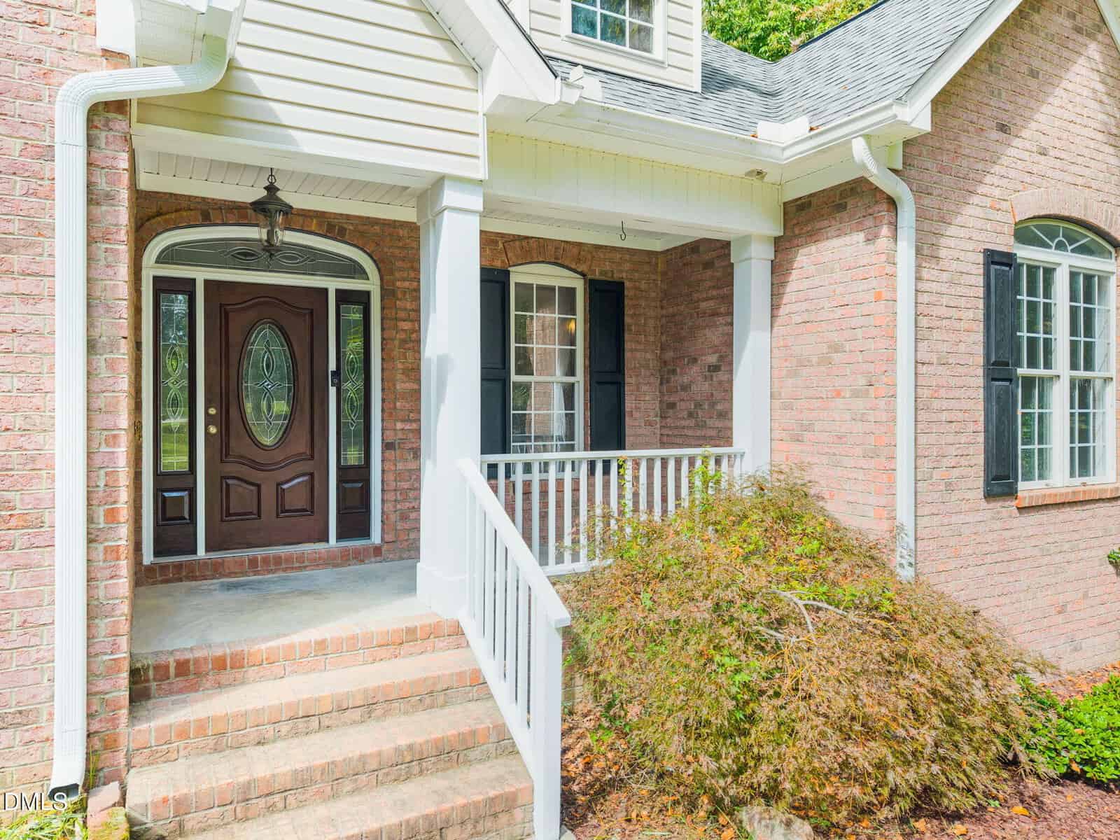 A brick house features a small front porch with white railings and columns, a dark wood front door with decorative glass, black shutters on the windows, and neatly trimmed shrubs beside the steps.