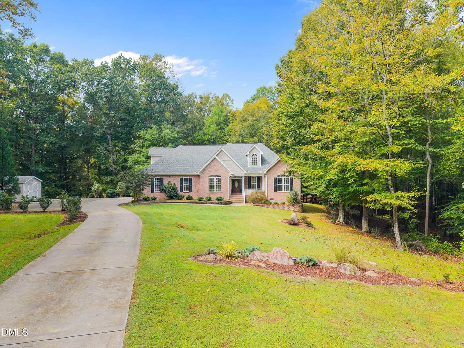 A single-story brick house with dormer windows sits at the end of a curved driveway, surrounded by a large green lawn and trees under a blue sky. A small shed is visible in the background to the left.
