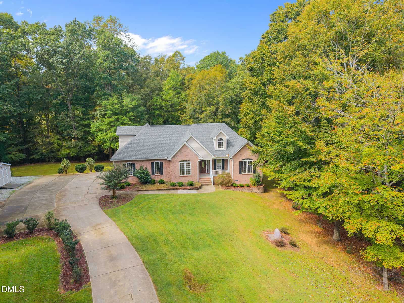 A brick house with a gray roof sits on a large, well-kept lawn surrounded by trees. The driveway curves to the left, leading to an attached garage. The sky is clear and blue.