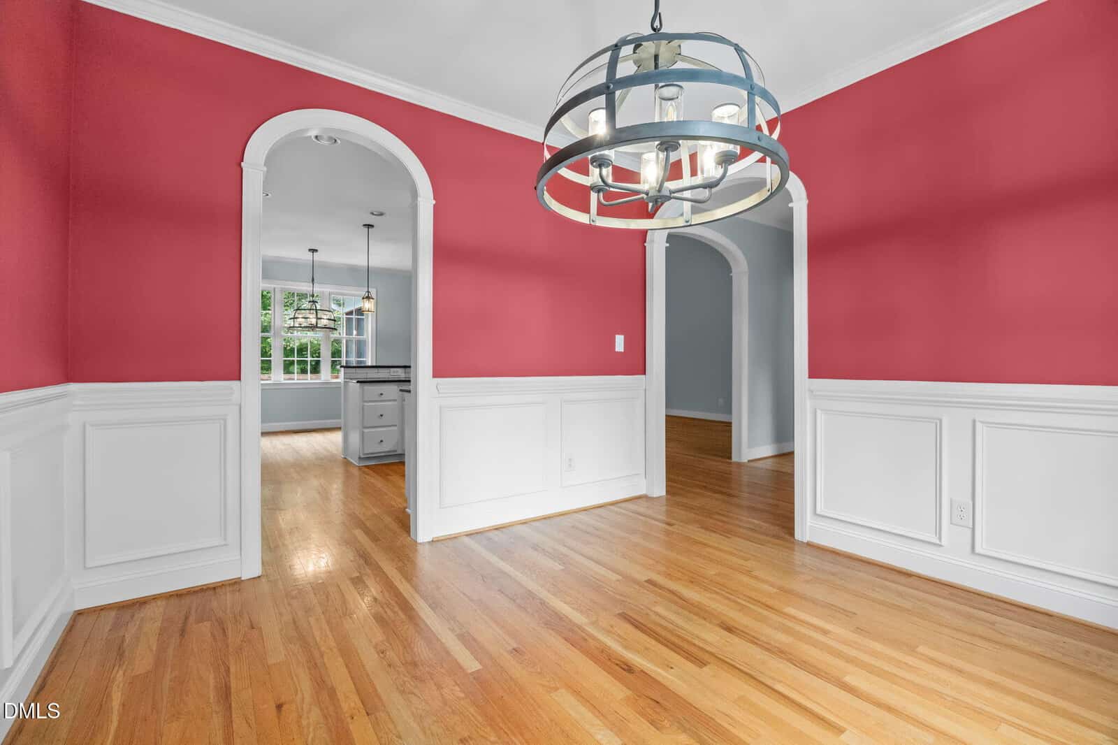 Dining room with red upper walls, white wainscoting, light hardwood floors, a modern circular chandelier, and two arched doorways leading to a kitchen and another room. Natural light from the kitchen window.