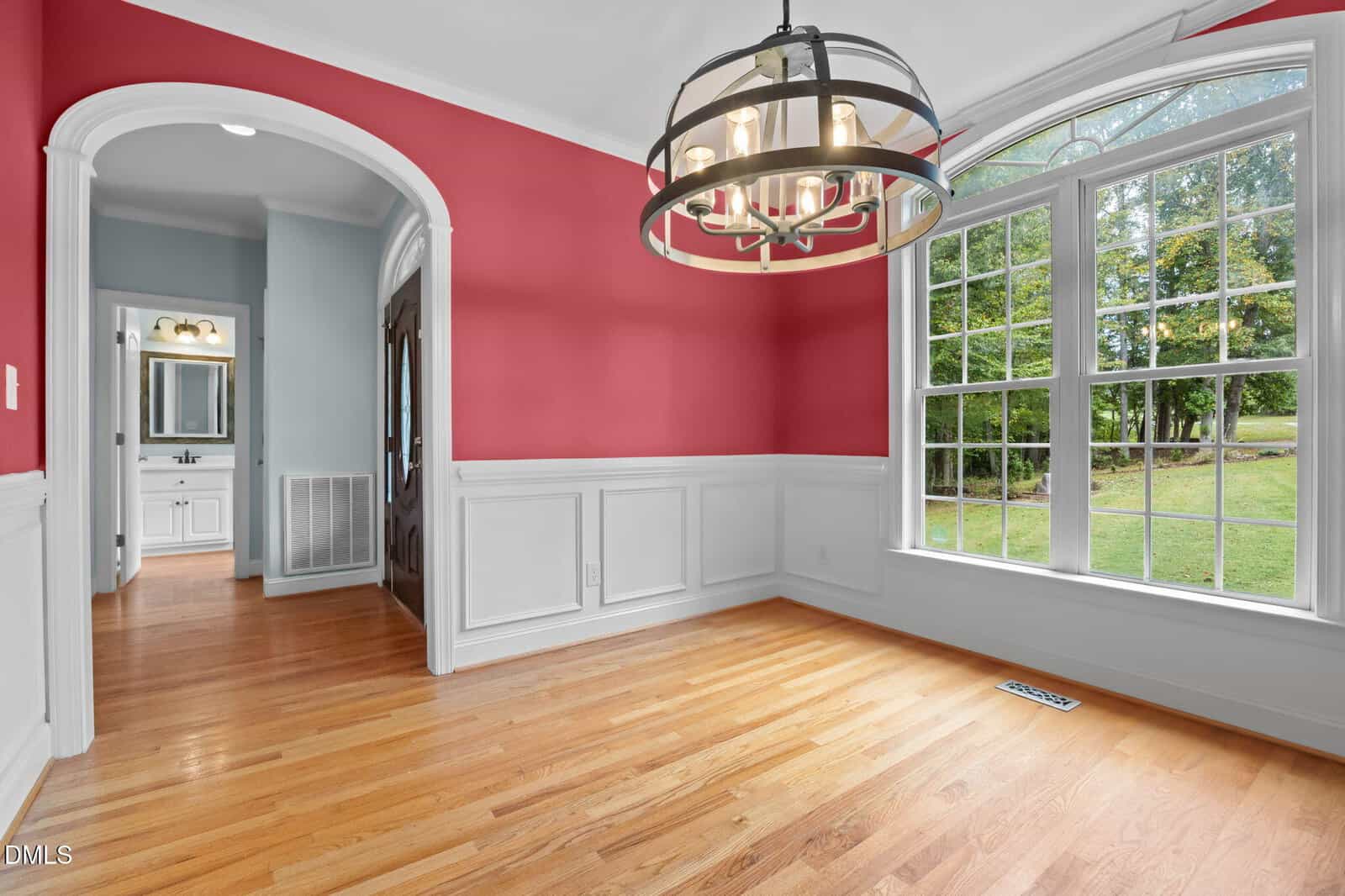 Dining room with red upper walls, white wainscoting, large arched window showing a green yard, wooden floor, modern chandelier, and an open doorway leading to a bathroom with a vanity and mirror.