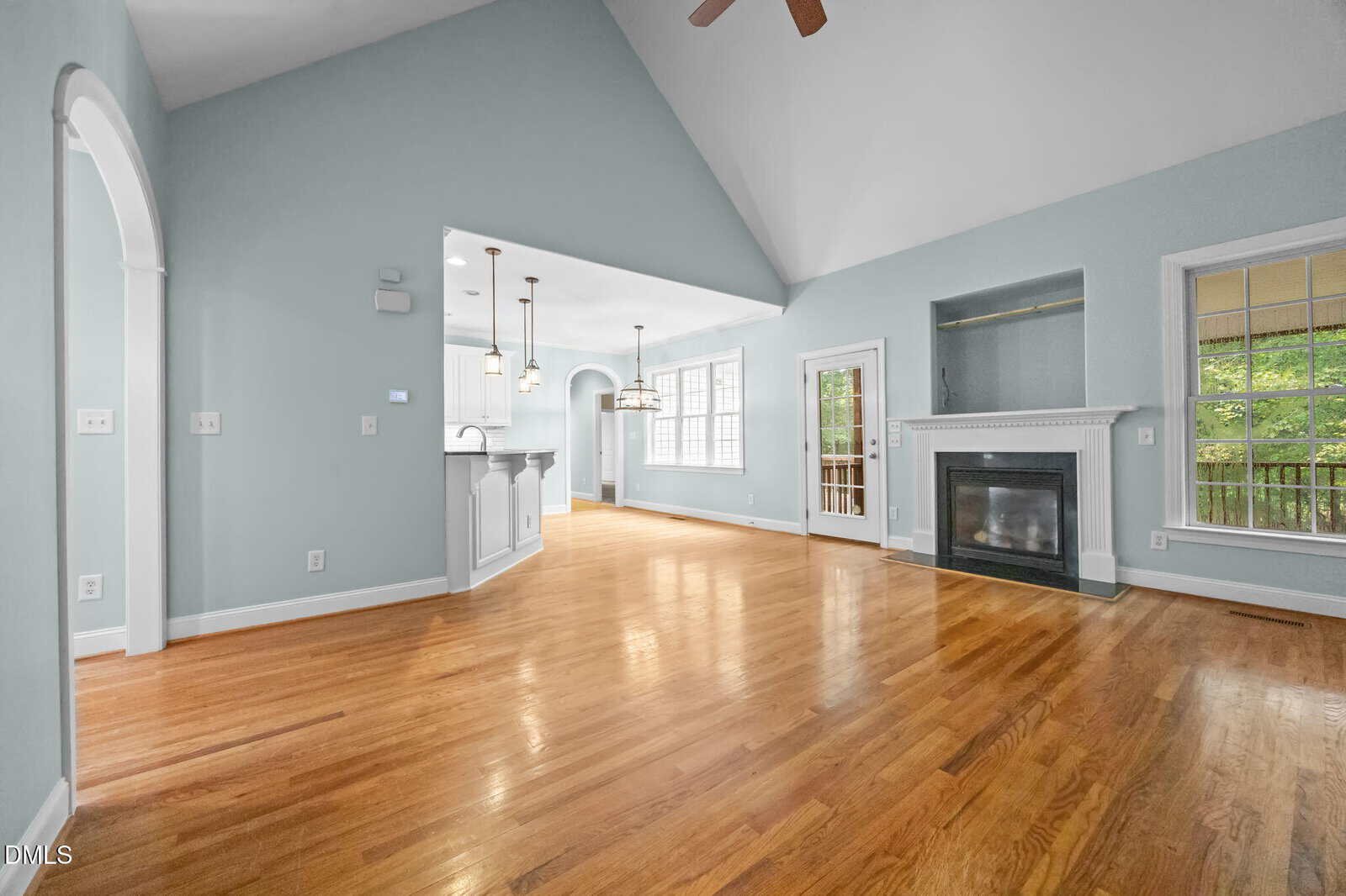 A spacious living area with light blue walls, vaulted ceiling, hardwood floors, a white fireplace, and large windows. An open kitchen with pendant lights and an arched entryway is visible to the left.