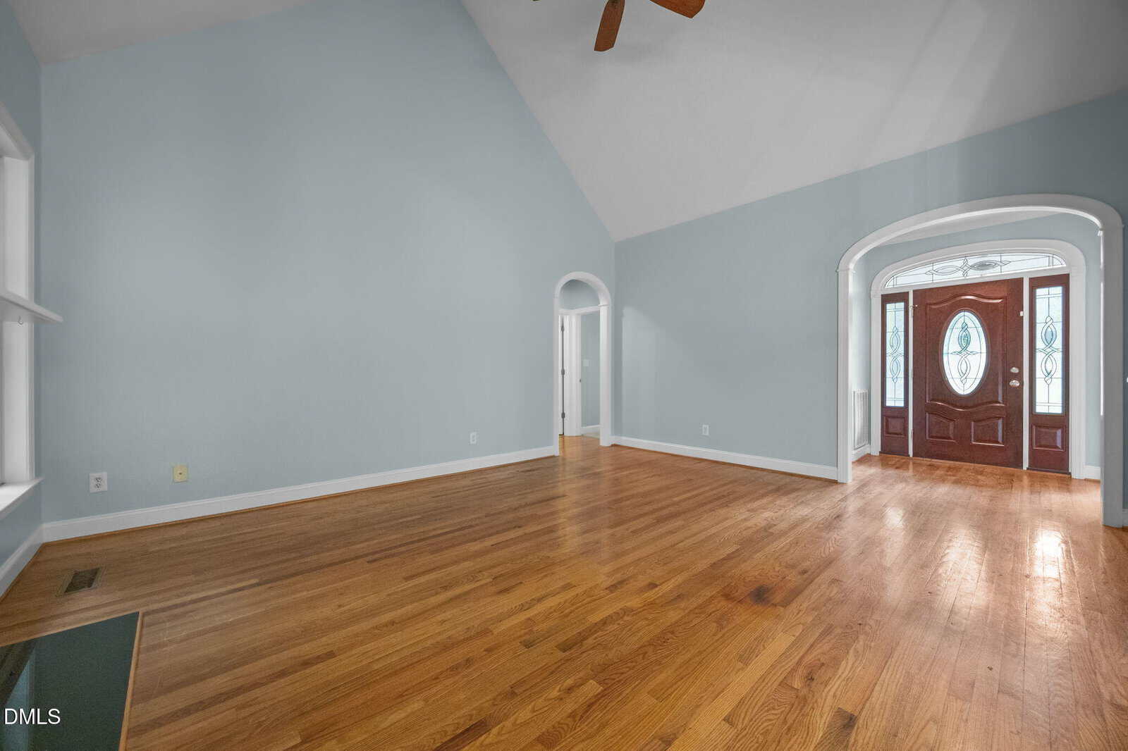 A spacious, empty living room with light blue walls, hardwood floors, a vaulted ceiling with a ceiling fan, and an arched entryway leading to a double front door with decorative glass panels.