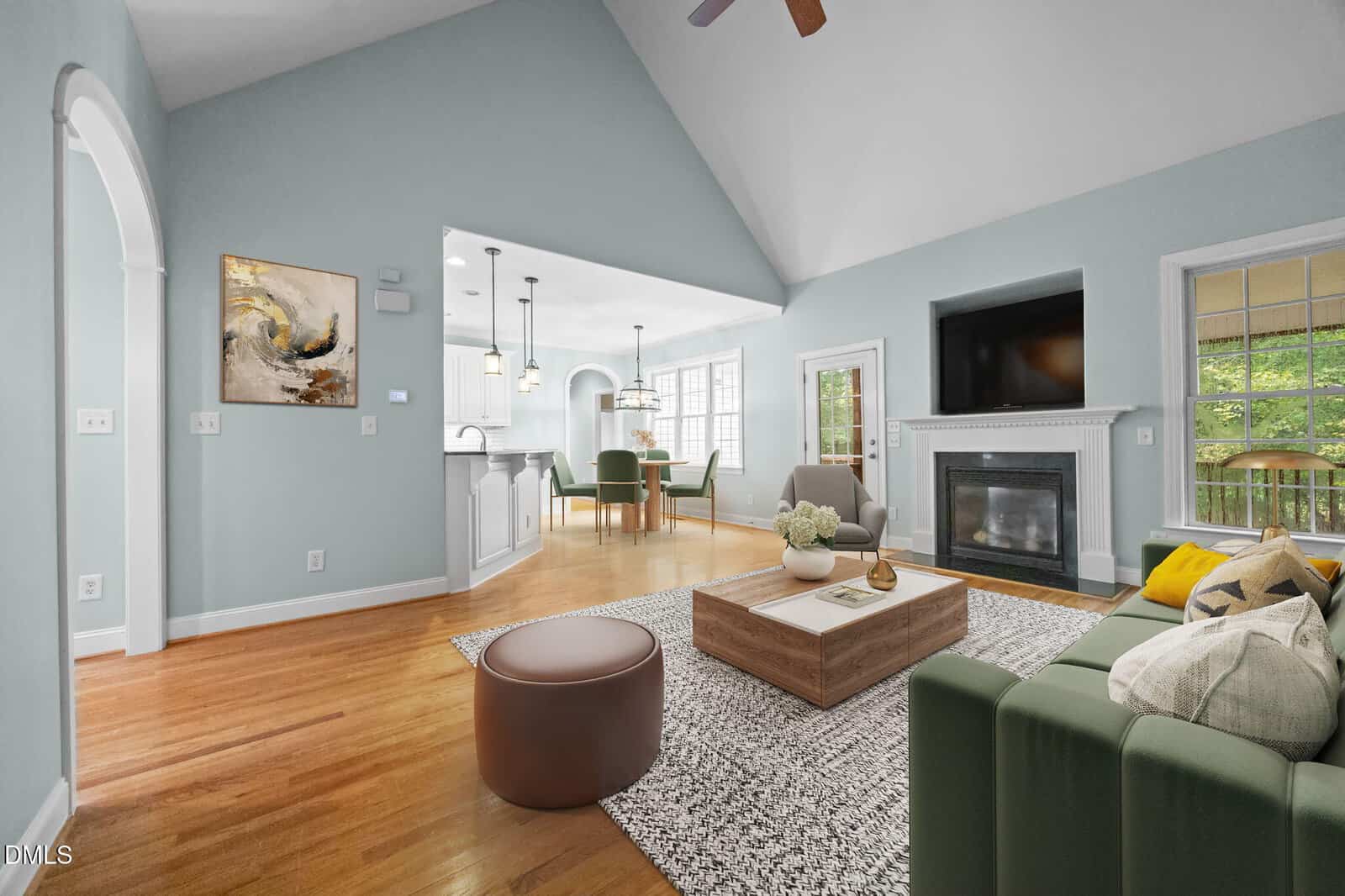 A modern living room with light blue walls, hardwood floors, a green sofa, patterned rug, coffee table, fireplace, abstract art, and open view into a bright kitchen and dining area with large windows.