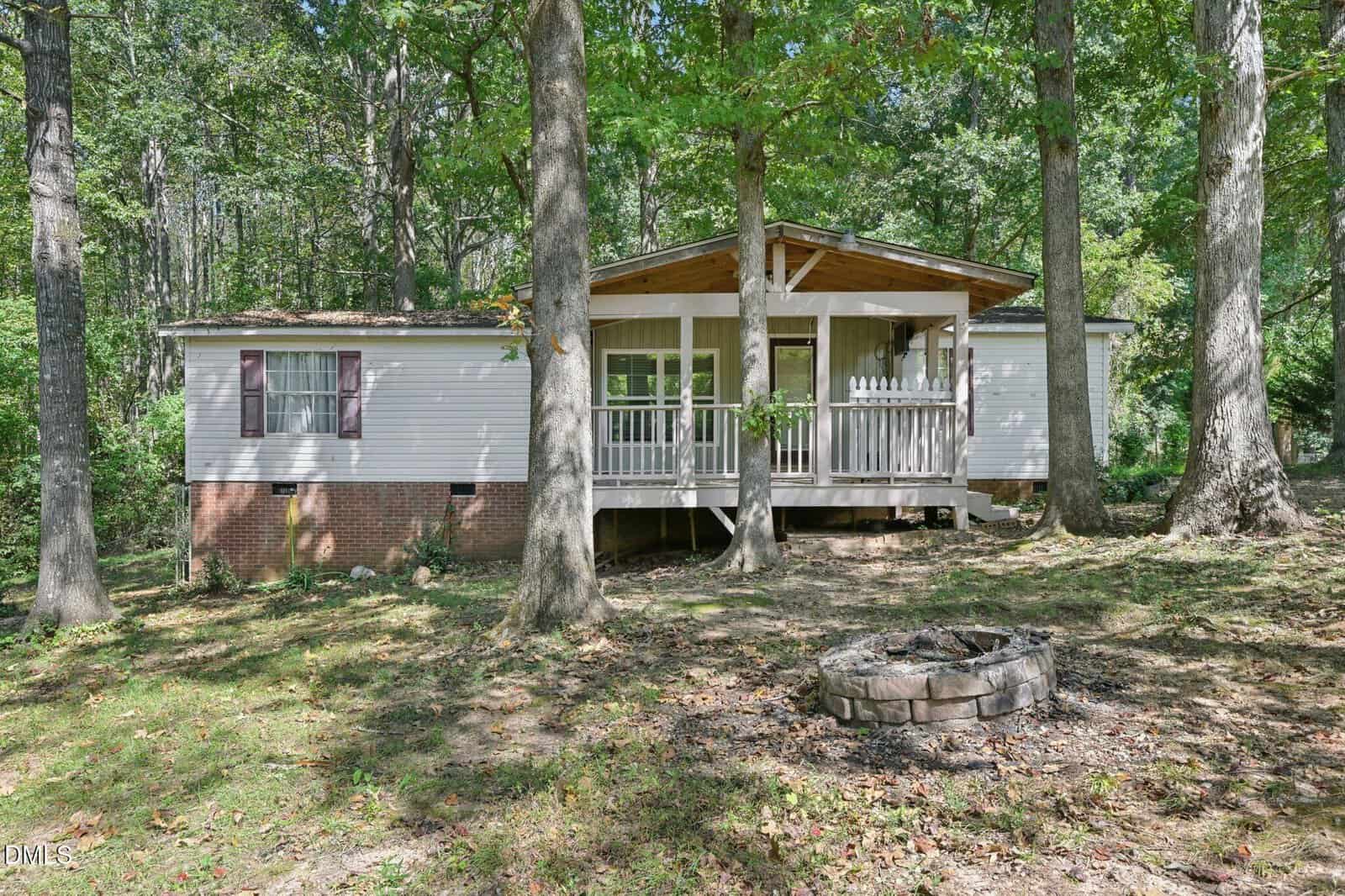 A white manufactured home with a front porch and steps, set on a brick foundation, is surrounded by tall trees at 12798 Farm Ridge Road in Zebulon. A small circular stone fire pit is in the grassy yard in front of the house.