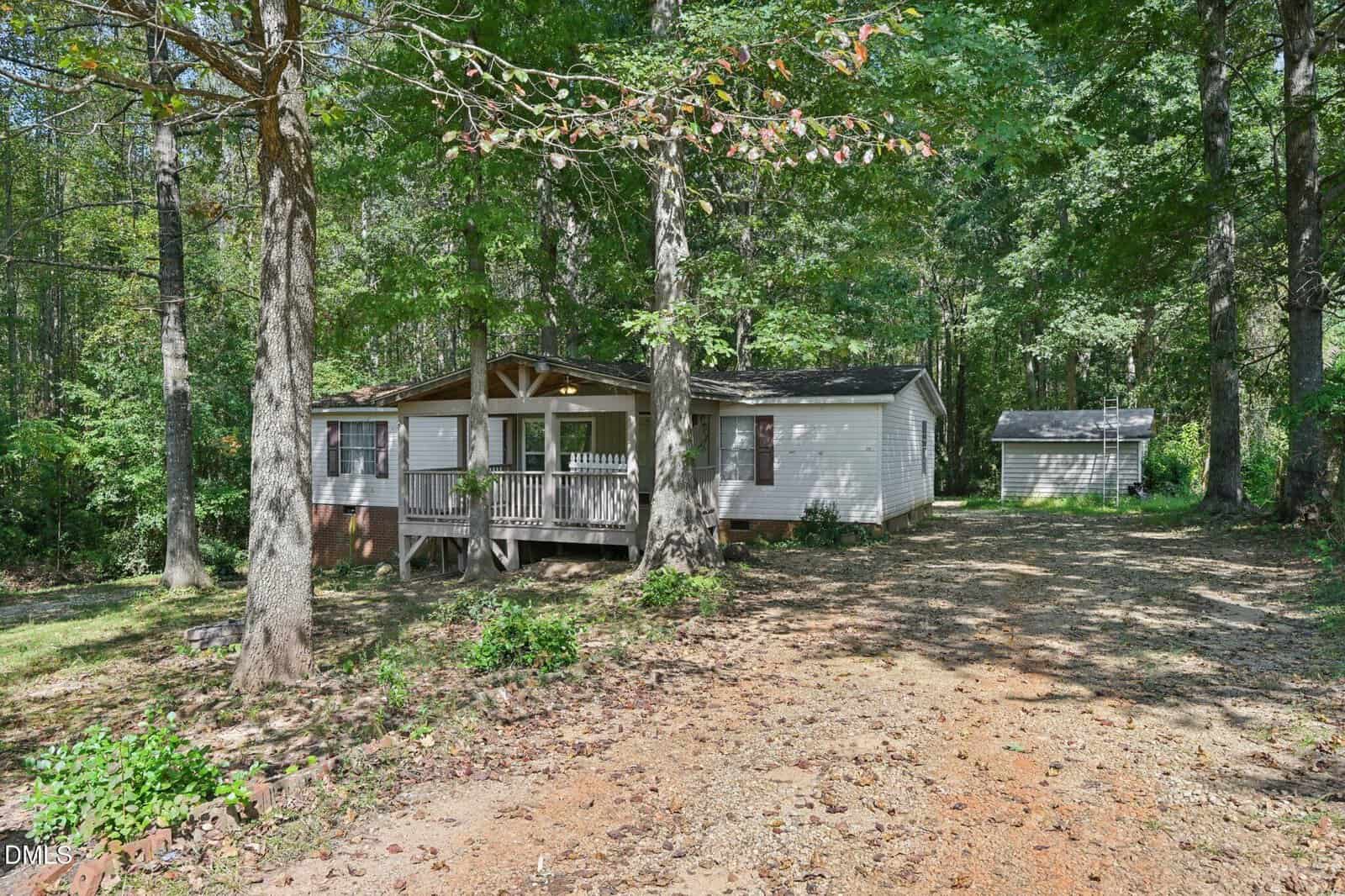 A single-story white house with a small front porch sits among tall trees on a dirt and gravel driveway. A detached shed or garage is visible in the background to the right. The area is shaded and surrounded by woods.