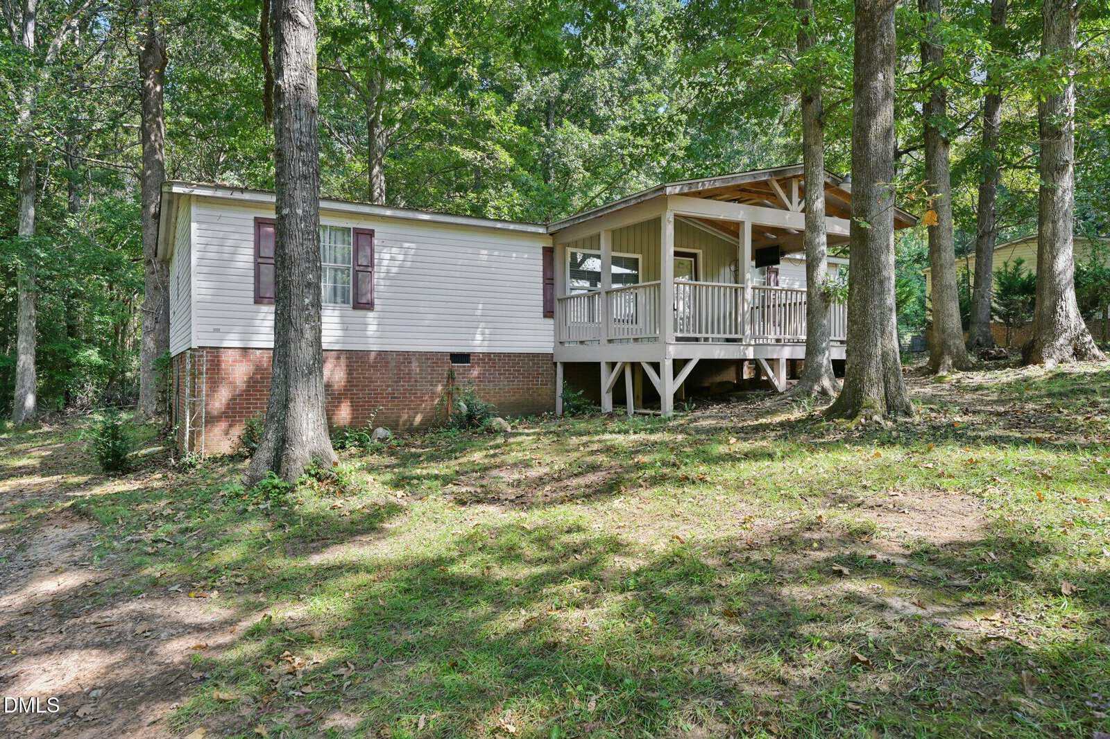Single-story house with white siding and red shutters, a brick foundation, and a covered front porch with railings. Surrounded by tall trees and set on a sloped grassy yard.