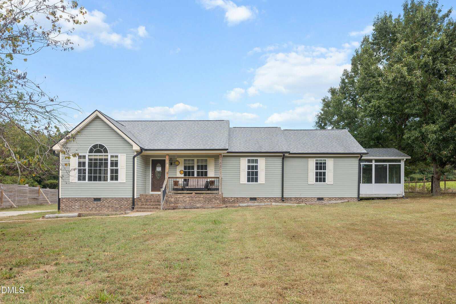 Single-story house with gray siding, brick foundation, front porch, and a screened-in room on the right. Located at 7397 Pearces Road in Louisburg, this home is surrounded by a large grassy yard and mature trees under a partly cloudy sky.