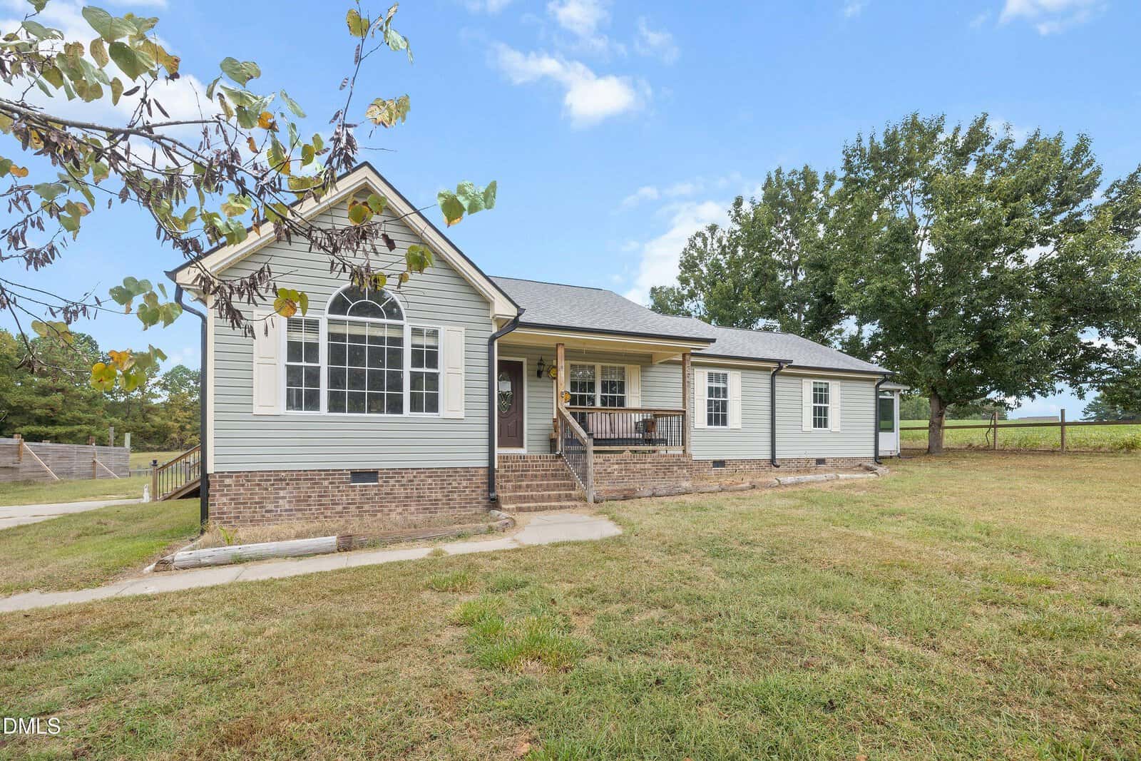 Single-story house with light gray siding, a brick foundation, and a small porch with steps. The yard is grassy with a large tree and a wooden fence in the background under a blue sky with scattered clouds.