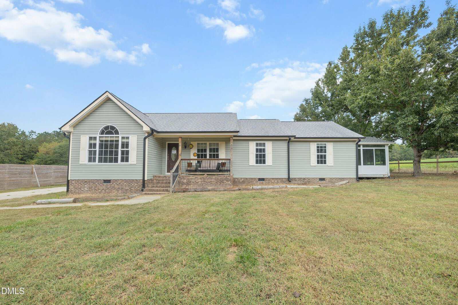 Single-story gray house with a brick foundation, covered front porch, and a screened-in side area, set on a grassy lawn with a large tree to the right and a wooden fence on the left under a blue sky with clouds.
