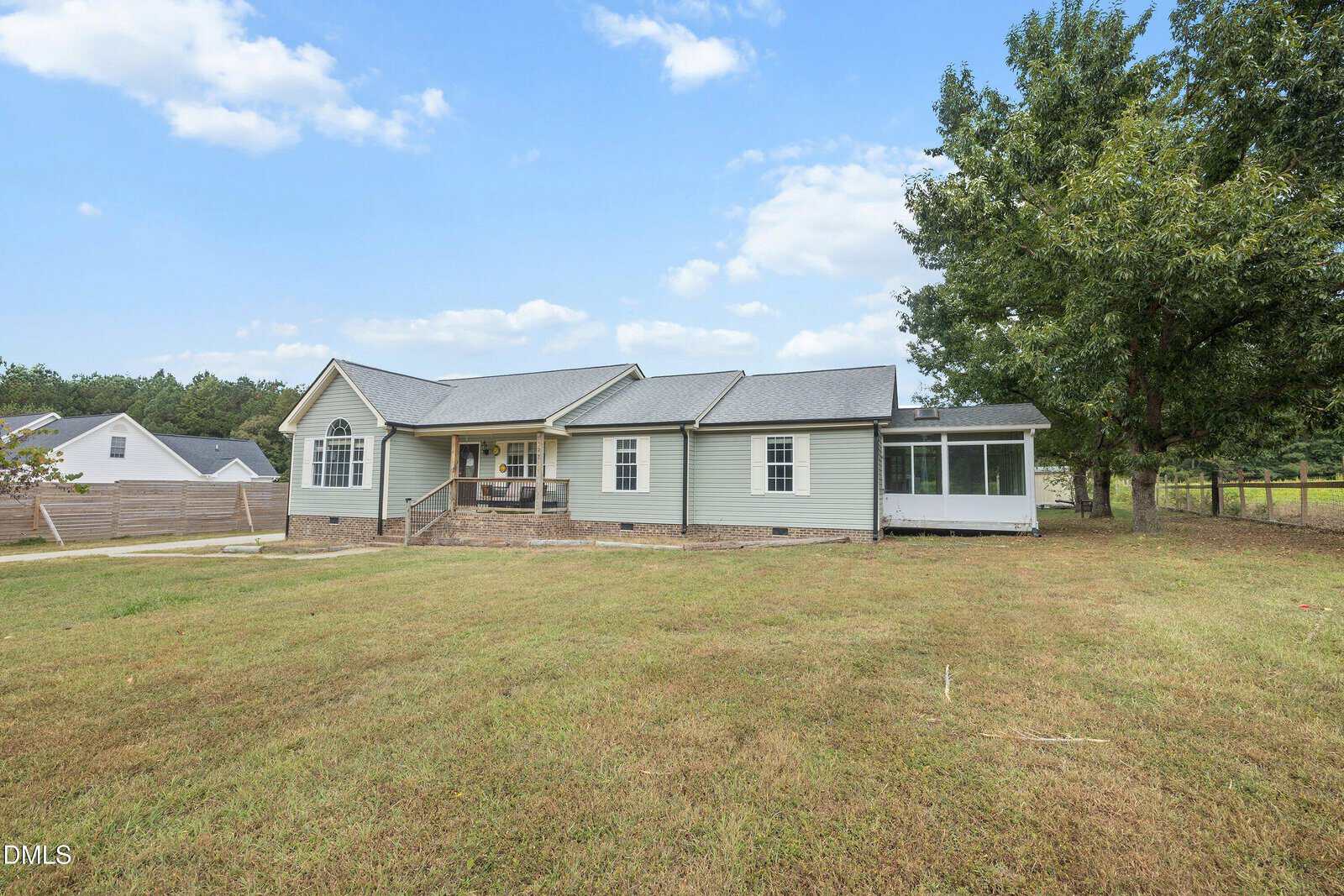 A single-story light green house with a covered front porch, brick foundation, and a screened-in porch on the right, surrounded by a spacious, grassy yard and bordered by trees under a partly cloudy sky.