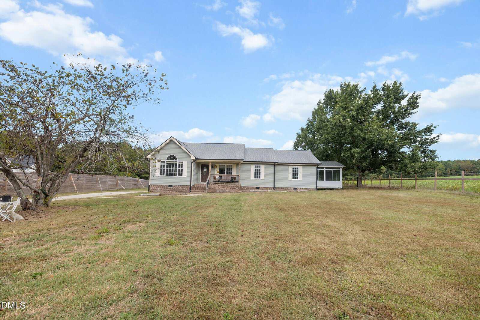 Single-story house with gray siding, gabled roof, and screened porch, set on a large grassy lot with minimal landscaping and trees in the background under a partly cloudy sky.