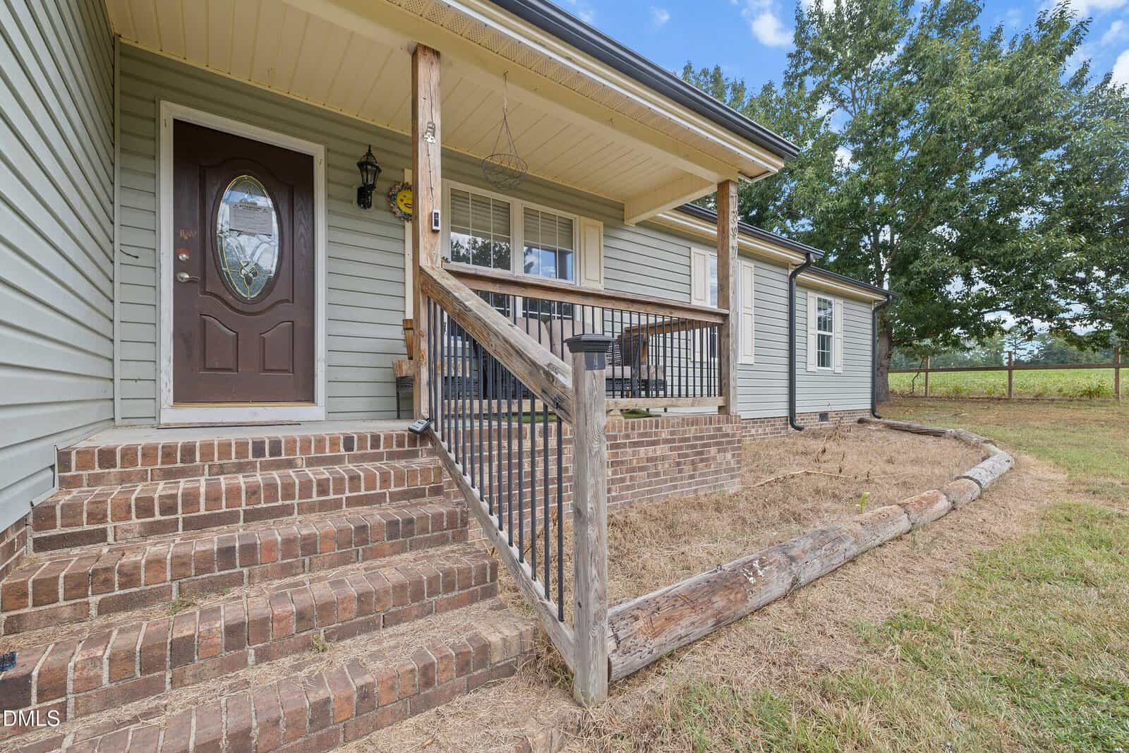 A front porch with brick steps, a black metal railing, and wooden posts attached to a house with gray siding and white trim. There is a brown front door with an oval window and a large yard with trees.
