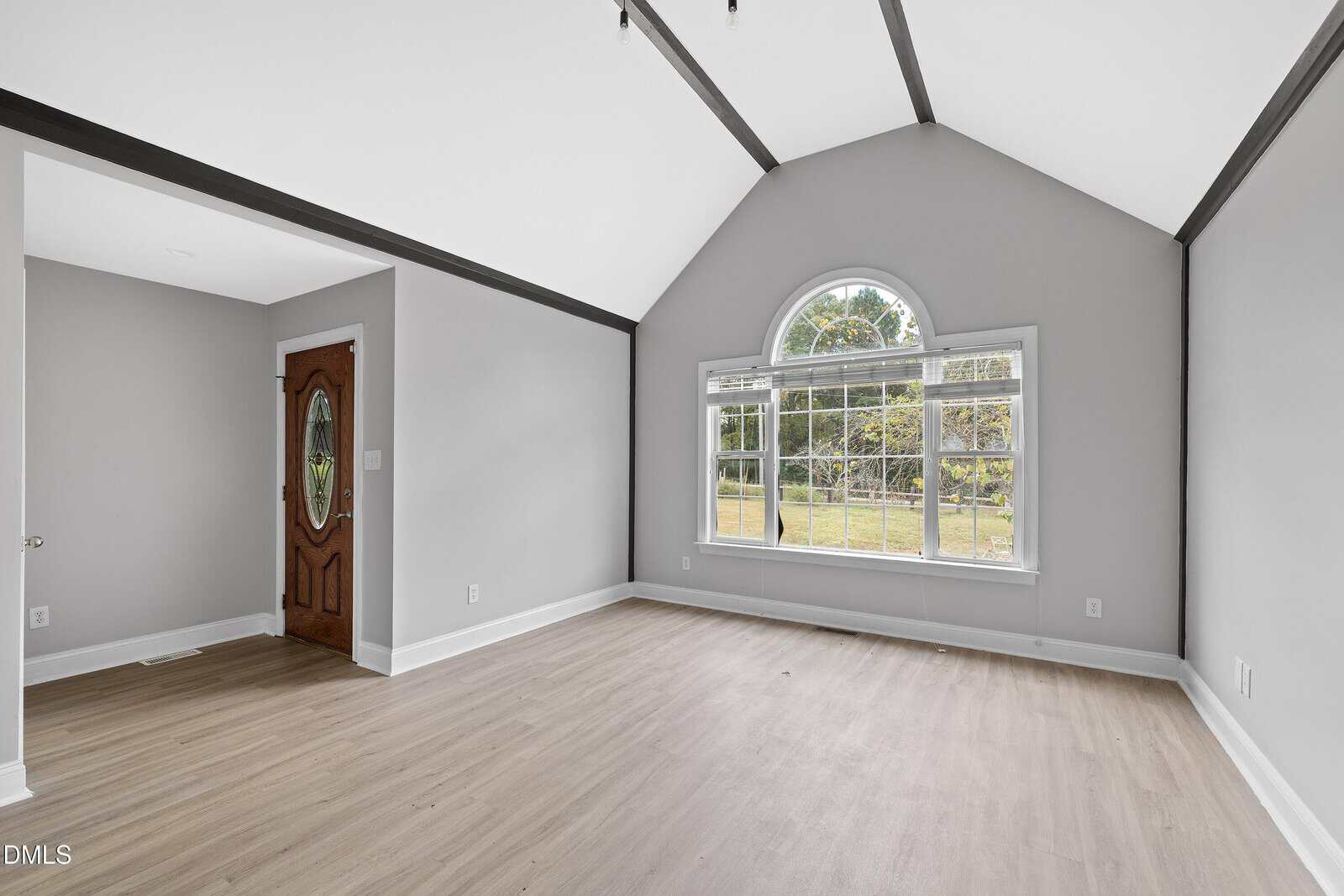 A bright, empty room with light wood flooring, gray walls, a vaulted ceiling with dark beams, a large arched window, and a wooden front door with an oval glass insert.
