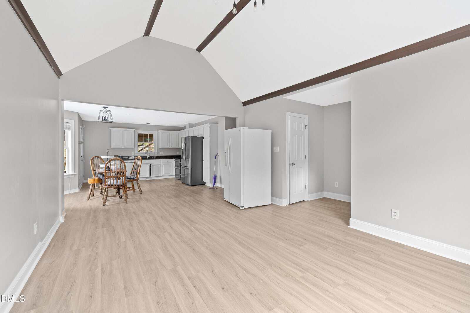 A spacious open-plan room with light wood flooring, vaulted ceiling with exposed beams, and neutral walls. The area leads to a kitchen with white cabinets, a dining table with chairs, and stainless steel appliances.