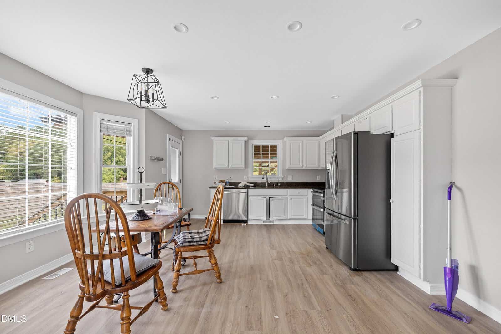 A kitchen with white cabinets, stainless steel appliances, light wood flooring, and a round dining table with four wooden chairs. A window and door let in natural light. A purple mop leans against the wall on the right.
