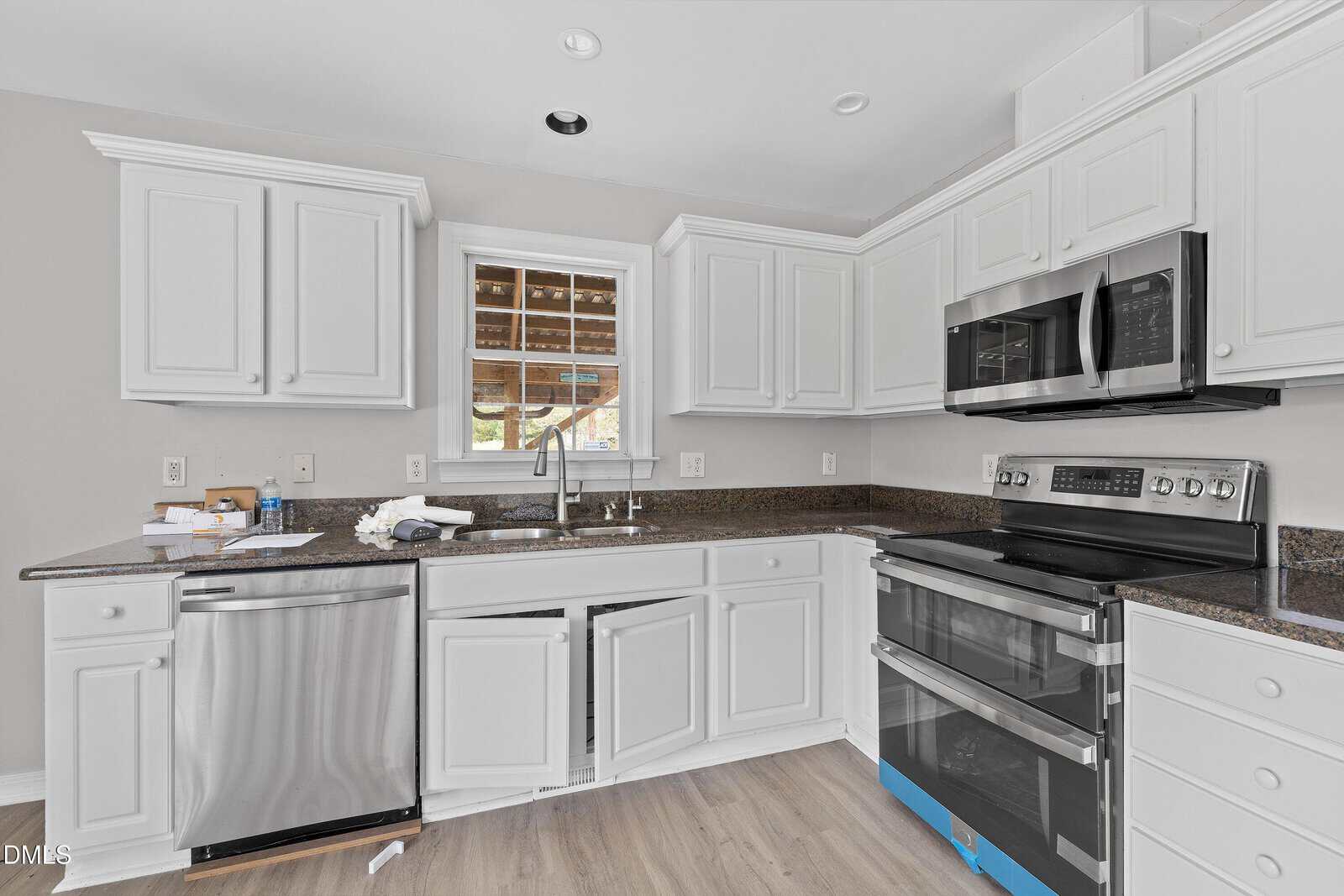 A modern kitchen with white cabinets, dark granite countertops, a stainless steel dishwasher, and a double oven. One cabinet and the dishwasher door are open. A window above the sink shows an outdoor view.