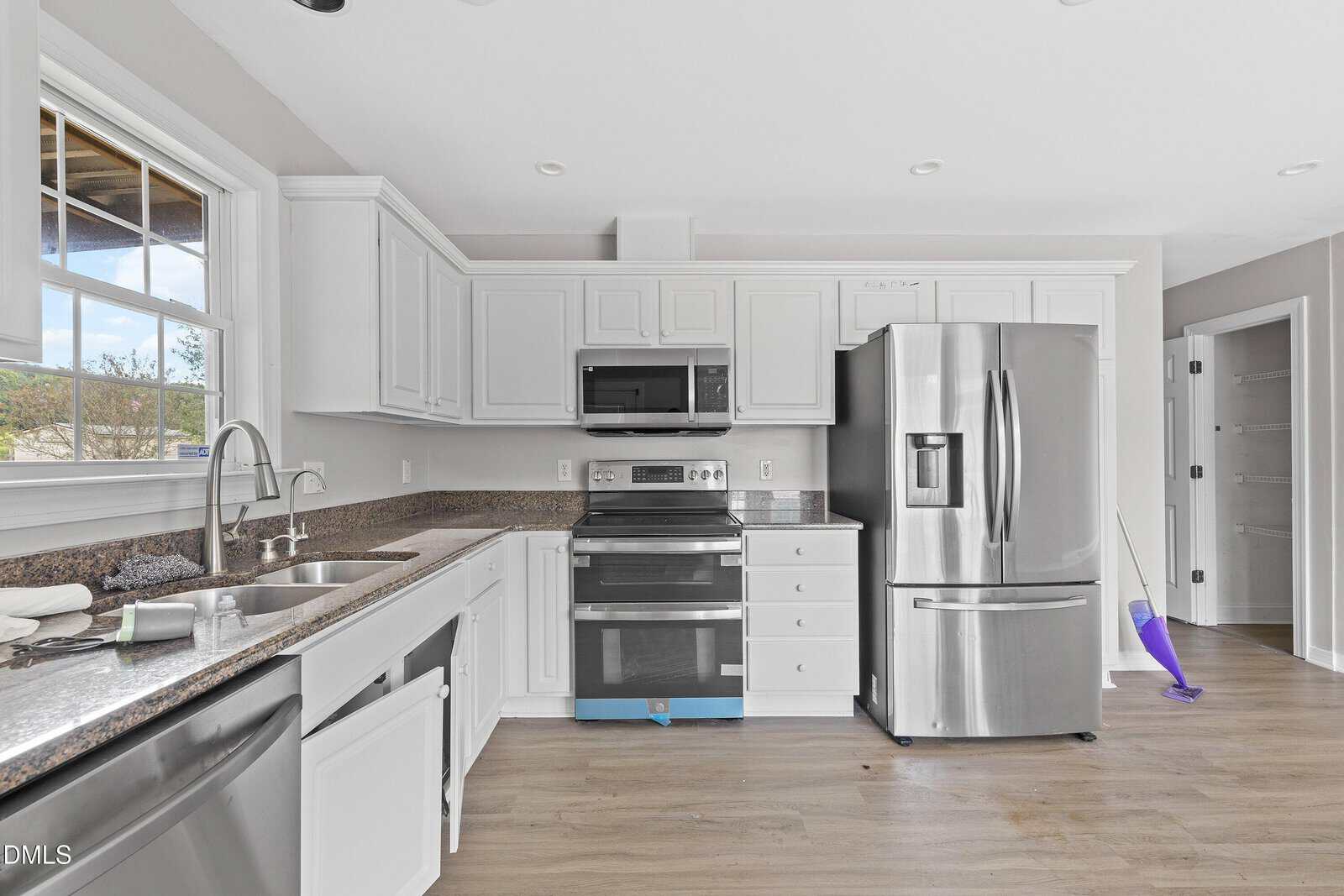 Modern kitchen with white cabinets, stainless steel appliances, a double sink under a window, and a refrigerator next to a broom and dustpan. Light wood flooring and neutral-colored walls complete the space.