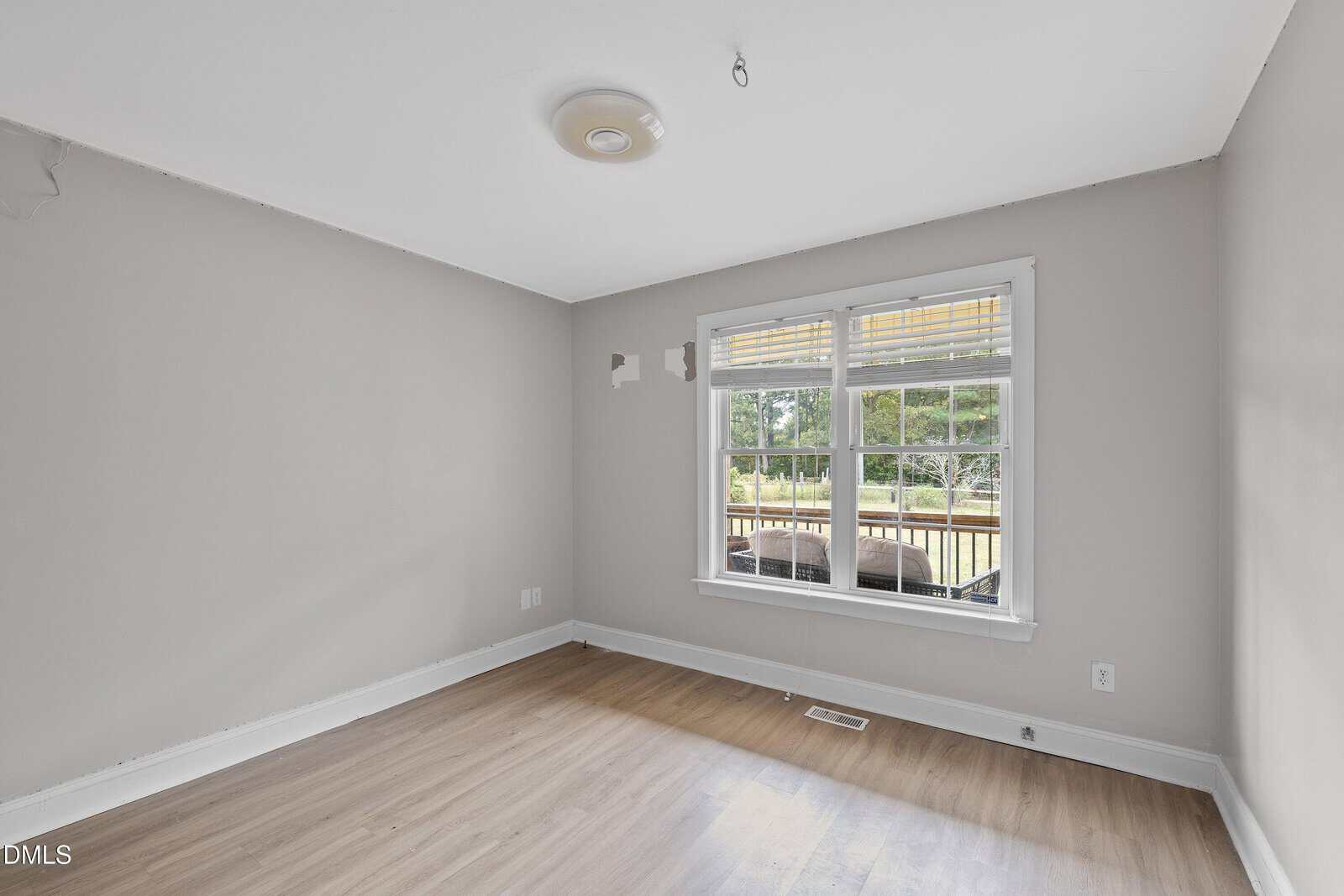 Empty room with light gray walls, light wood flooring, a ceiling light fixture, two wall outlets, and a large window with blinds, showing a wooden deck and greenery outside.