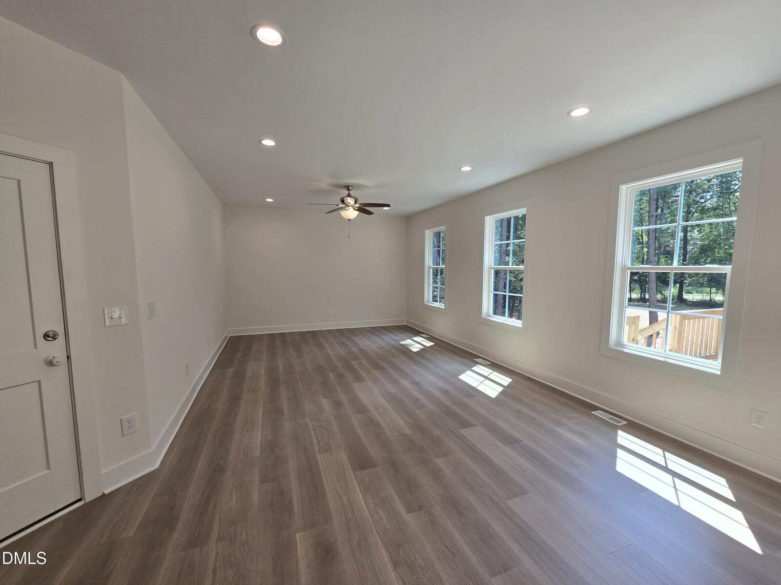 A spacious, empty room with light-colored wood flooring, white walls, recessed ceiling lights, a ceiling fan, and several large windows letting in natural light.