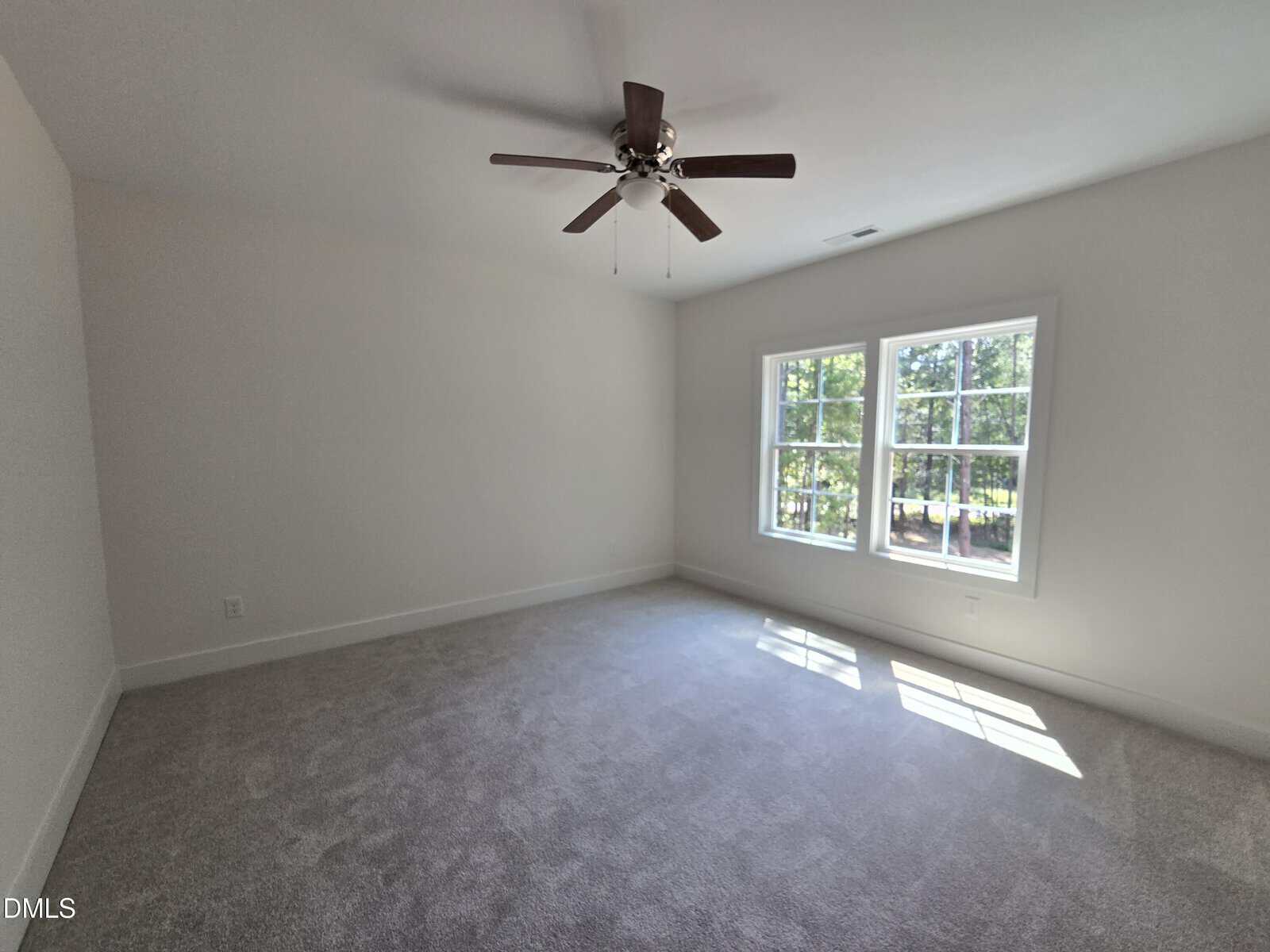 Empty room with light-colored carpet, beige walls, a ceiling fan with wooden blades, and a large window letting in sunlight. Trees and greenery are visible outside the window.
