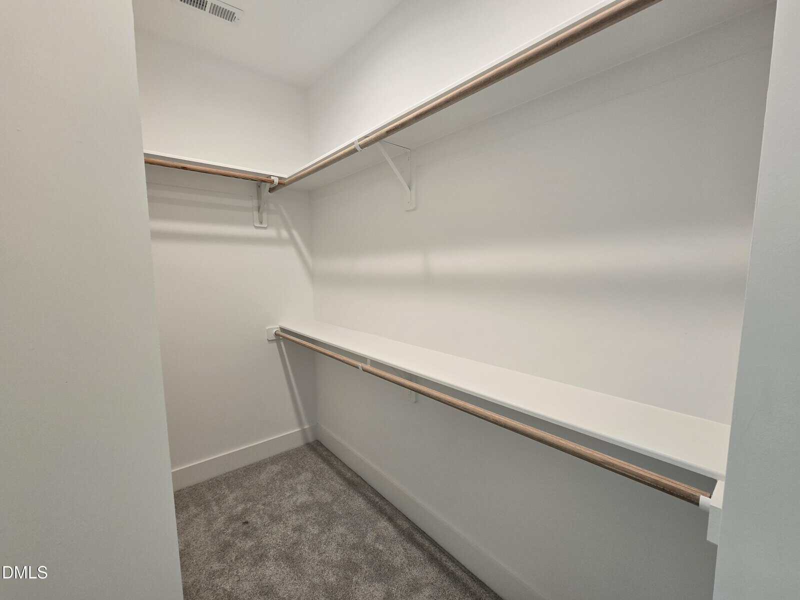 A spacious, empty walk-in closet with white walls, carpeted floor, and wooden hanging rods on both sides for clothes.
