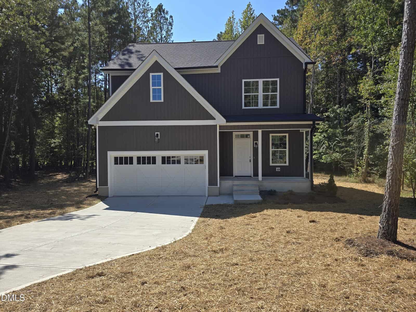 Two-story dark gray house with a white garage door, white trim, and front porch. The house sits on a curved concrete driveway surrounded by pine straw and trees in the background under a clear sky.