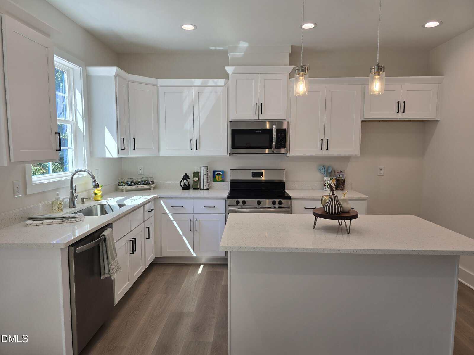 Modern kitchen with white cabinets, stainless steel appliances, a center island, pendant lights, and light wood flooring. A window above the sink lets in natural light and decorative items are placed on the counters.