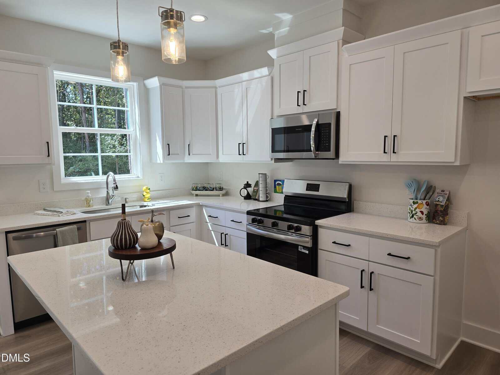 Modern kitchen with white cabinets, stainless steel appliances, a gas stove, microwave, and dishwasher. An island with a white quartz countertop sits in the center, decorated with a tray. A window lets in natural light.