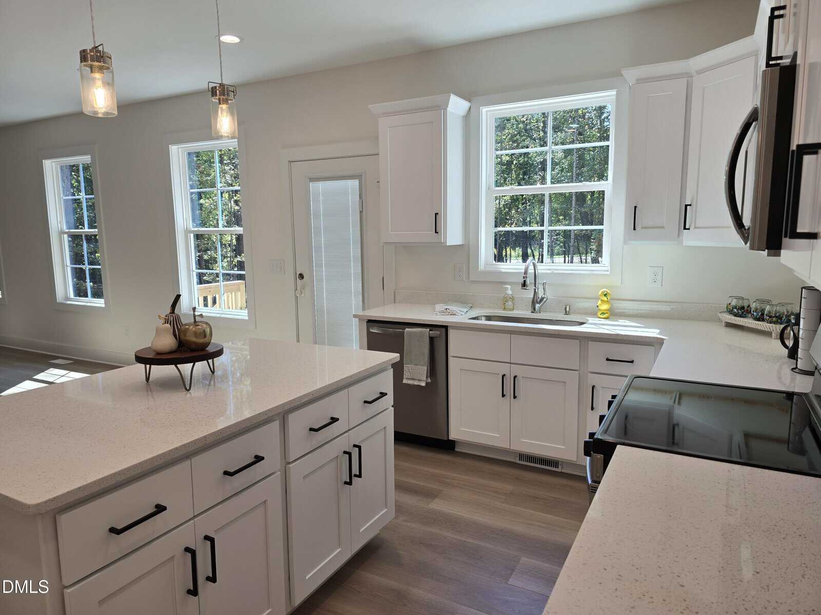 Bright modern kitchen with white cabinets, stainless steel appliances, a large island, quartz countertops, pendant lighting, and multiple windows letting in natural light. A small tray with decor is on the island.