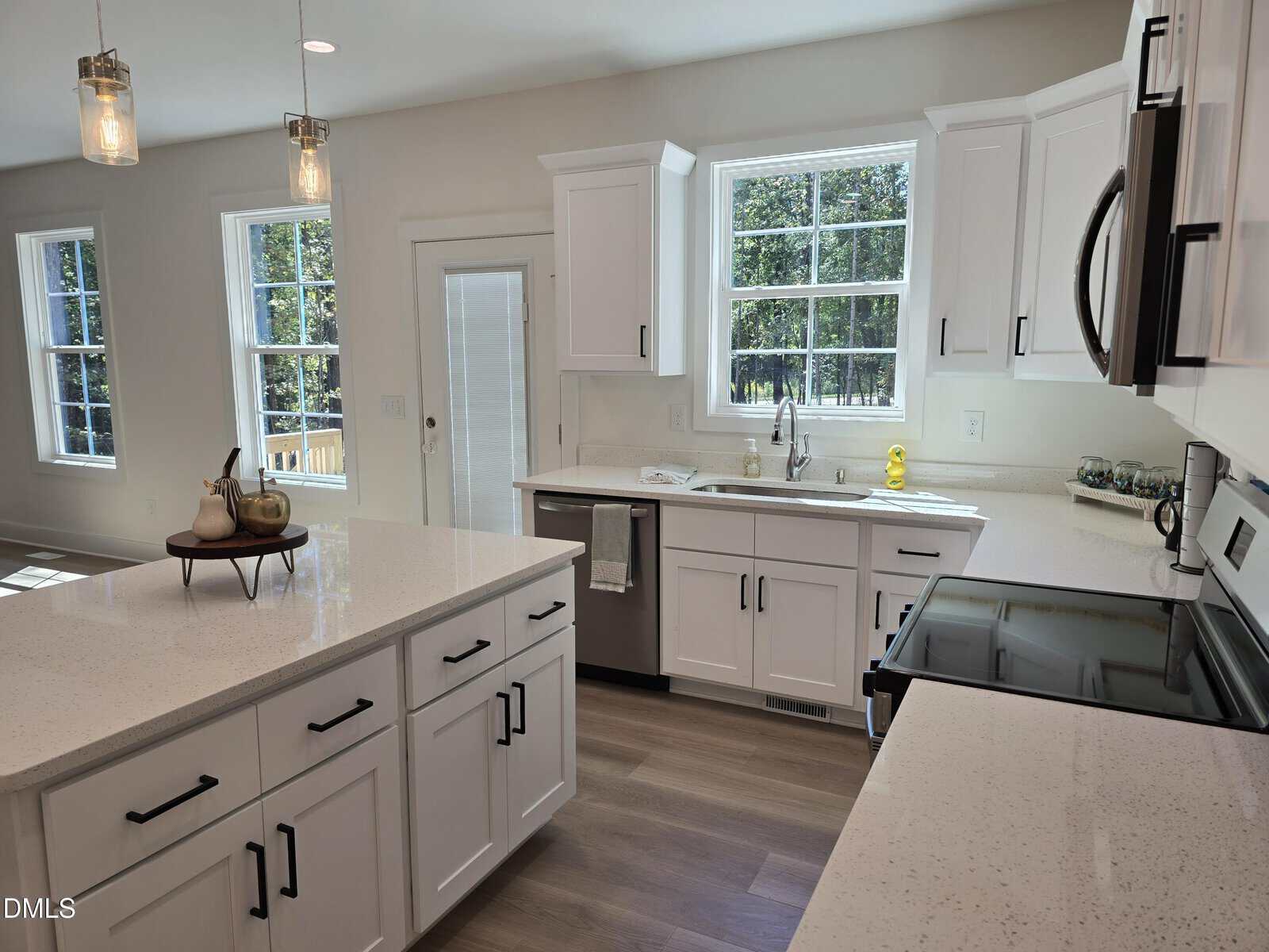 A modern kitchen with white cabinets, stainless steel appliances, a central island with fruit decor, large windows, and light-colored wood flooring, all well-lit by natural light.