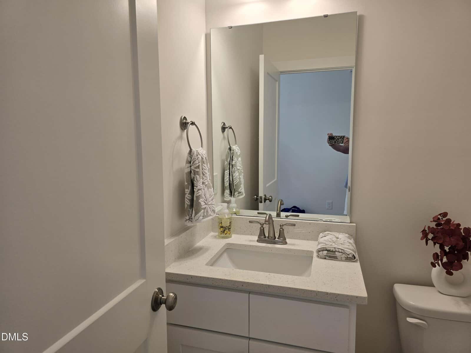 A small bathroom with a white vanity, silver faucet, and a rectangular mirror. A hand towel hangs on the wall, soap and folded towels are on the counter, and red flowers sit on the toilet tank. A door is partially open.