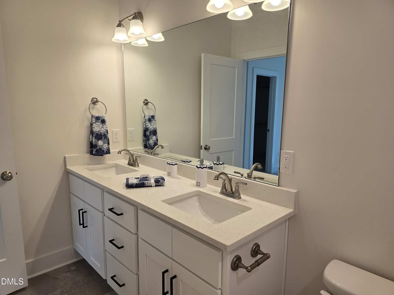 A modern bathroom features a large mirror above a double-sink vanity with white cabinets, chrome fixtures, two towel rings with blue-patterned towels, and overhead lighting. A partially open door is visible in the background.