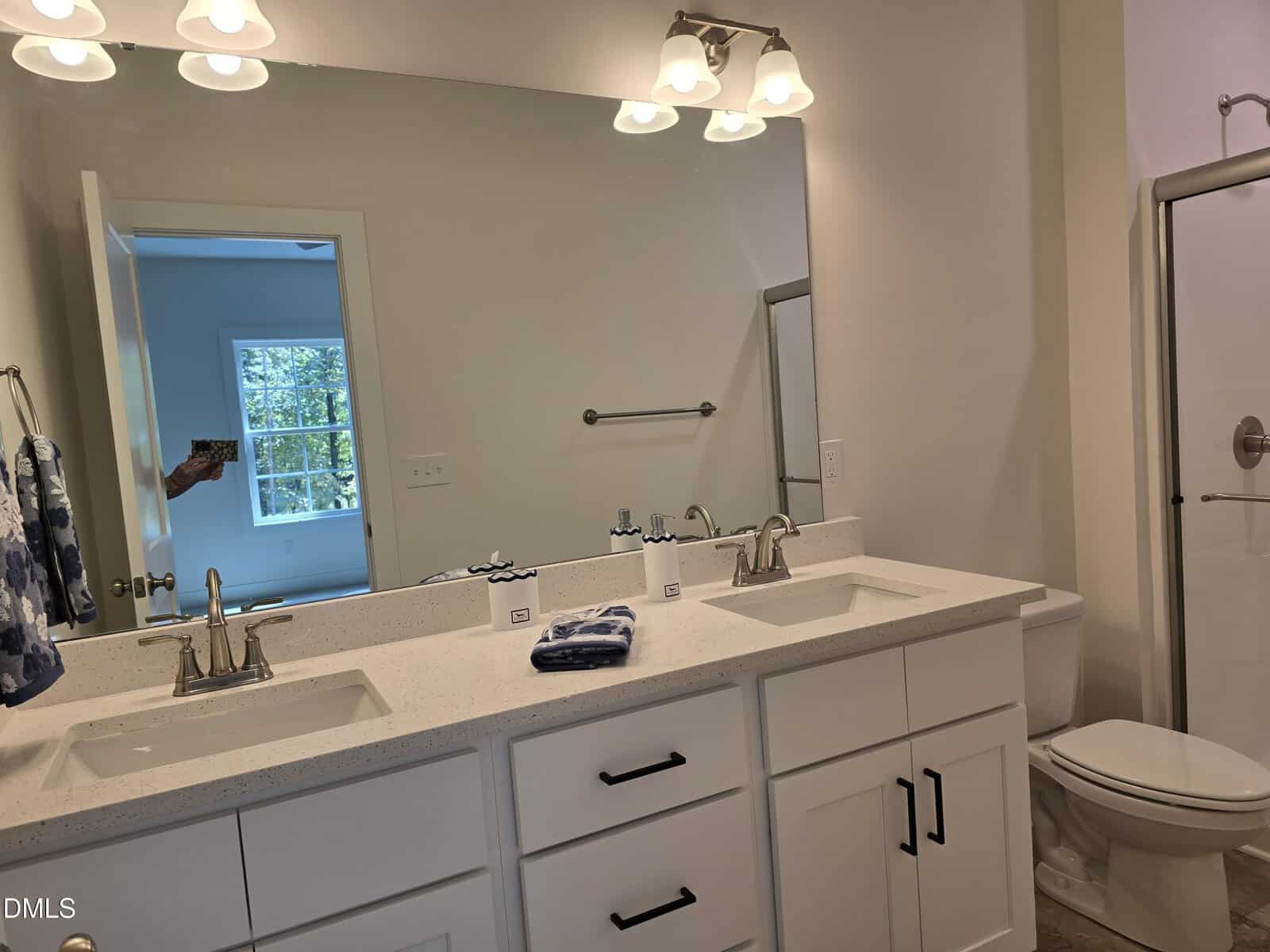 A bathroom with a double sink vanity, white cabinets, a large mirror, overhead lights, a white countertop, a towel rack, and a glass-enclosed shower. A window in an adjacent room is visible through an open door.