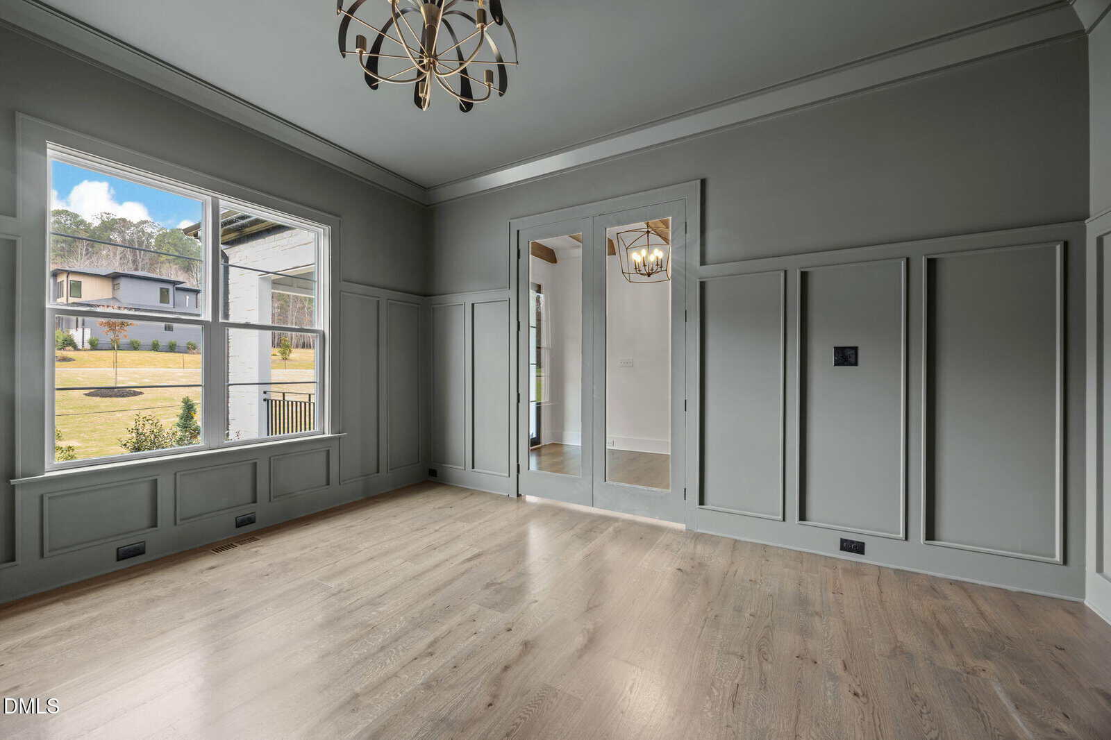 A spacious, empty room with light wood floors, gray paneled walls, a large window, glass double doors, and a modern chandelier hanging from the ceiling. Sunlight streams in through the window, brightening the space.