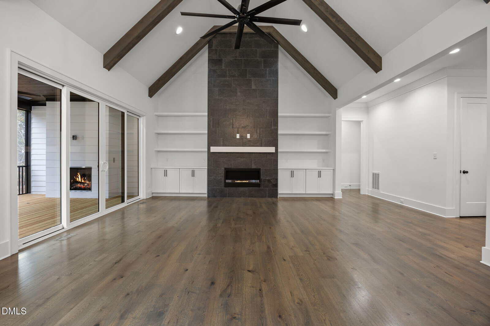 Modern living room with vaulted ceiling, dark accent fireplace, built-in shelves, and sliding glass doors opening to a covered patio with a fireplace. Hardwood floors and recessed lighting complete the space.
