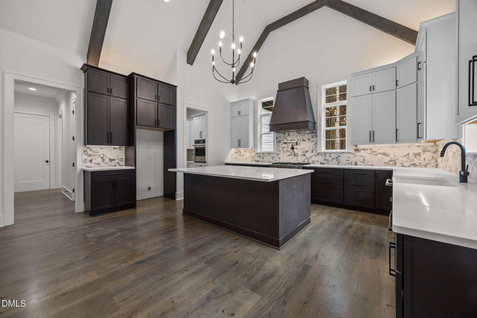 Modern kitchen with dark lower cabinets and island, light upper cabinets, marble backsplash, stainless steel appliances, wood flooring, exposed ceiling beams, a large chandelier, and high vaulted ceiling.
