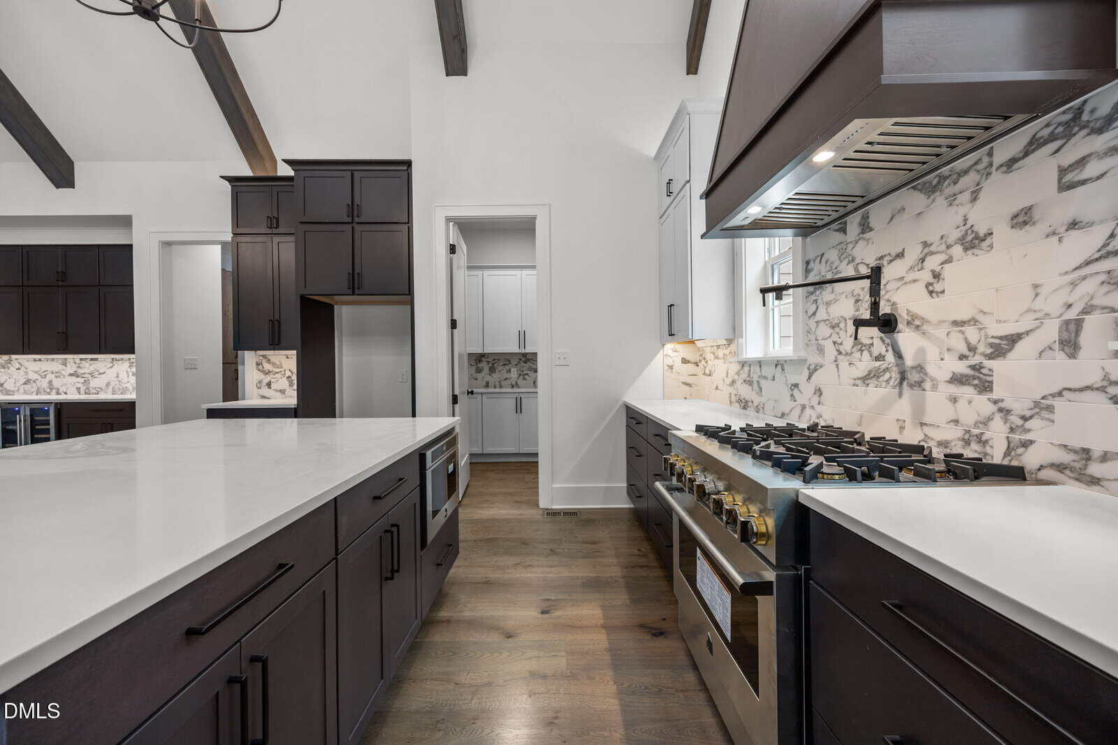 Modern kitchen with dark cabinets, white marble countertops, a large gas stove, and marble backsplash. The space features wood floors, exposed ceiling beams, and a view into an adjoining pantry area.
