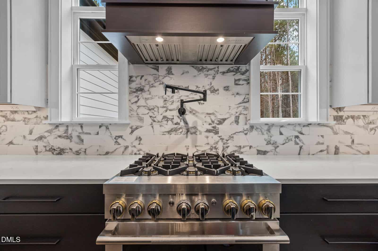 A modern stainless steel gas stove with six burners and gold-tone knobs is set against a white marble backsplash with gray veining, beneath a range hood and two windows on either side.