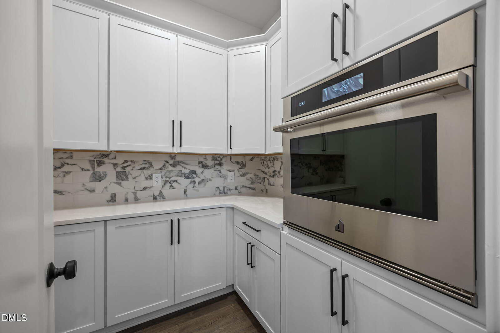 Modern kitchen with white cabinets, marble backsplash, and built-in stainless steel oven. Dark hardware contrasts with the white cabinetry, and wood floors add warmth to the space.
