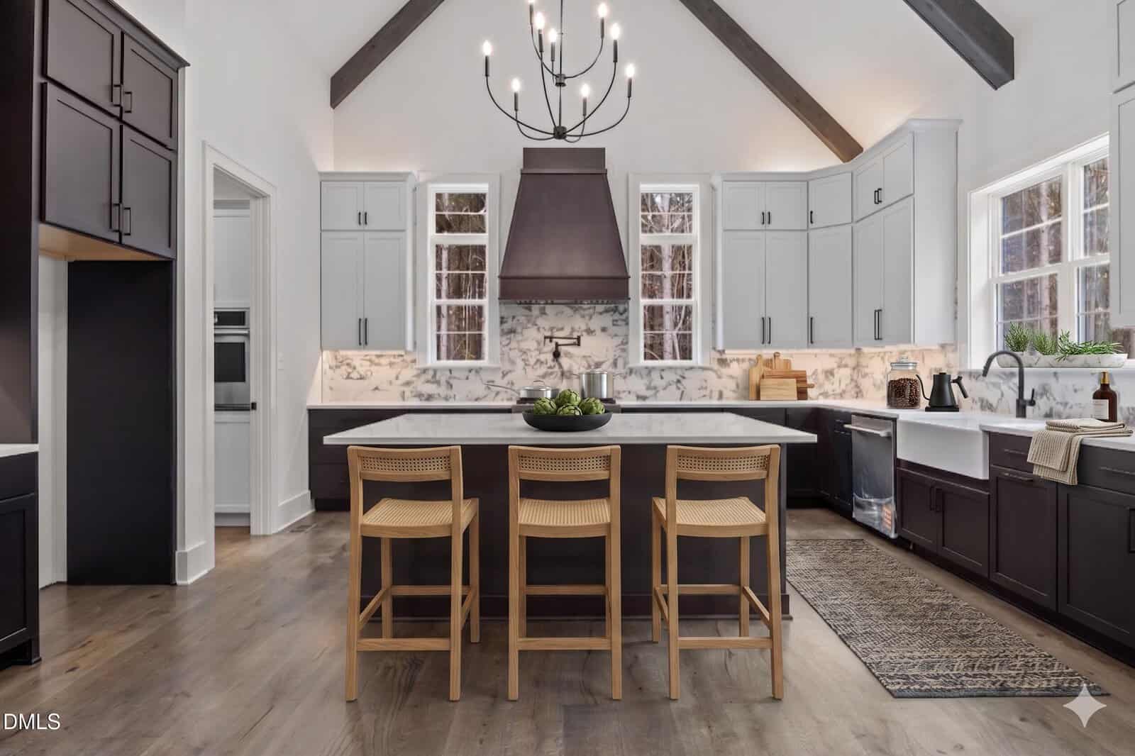 Modern kitchen with vaulted ceiling, dark wood beams, marble backsplash, white and dark cabinetry, large island with three woven chairs, farmhouse sink under windows, and a black chandelier above the island.