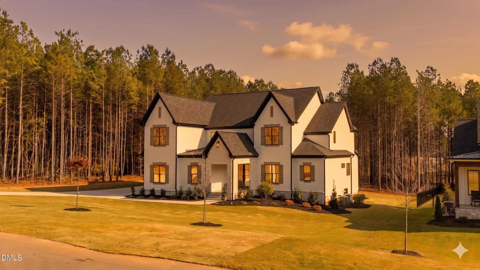 A large modern white house with black trim sits on a neatly manicured lawn, surrounded by tall pine trees under a warm, golden sky. The driveway curves beside the house and young trees are planted in the yard.