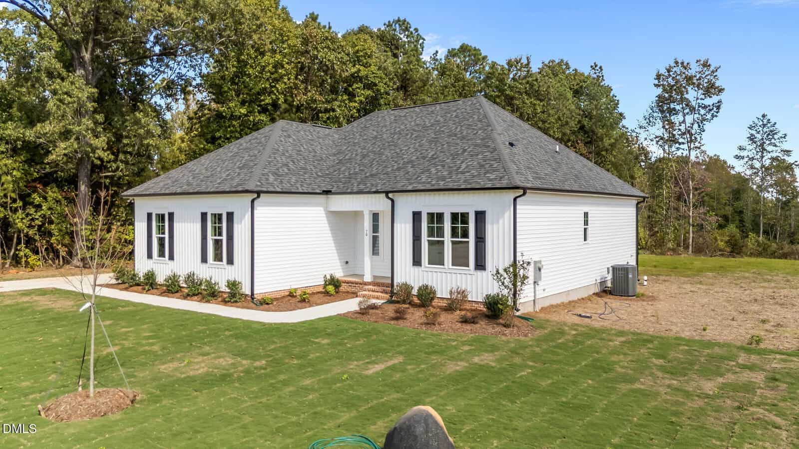 A single-story white house with black shutters and a dark gray roof sits on a newly landscaped lawn, surrounded by trees under a clear blue sky. There are new plants along the house and an air conditioning unit on the right side.