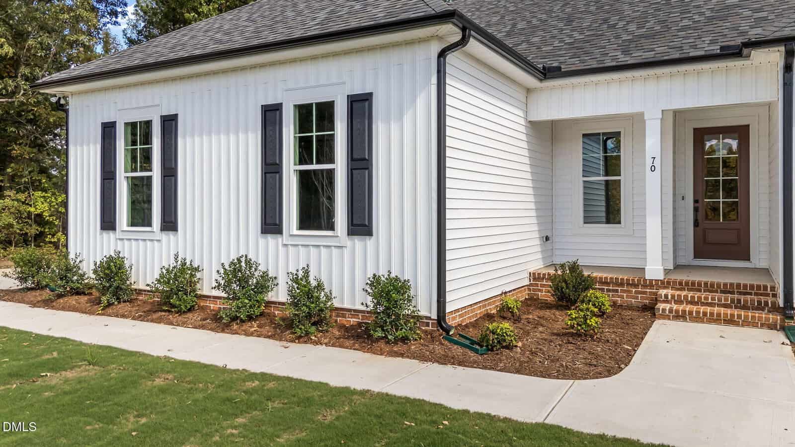 A white house with vertical siding, black shutters, and a brown front door, featuring three windows, a small covered porch with brick steps, and neatly landscaped bushes along a sidewalk.
