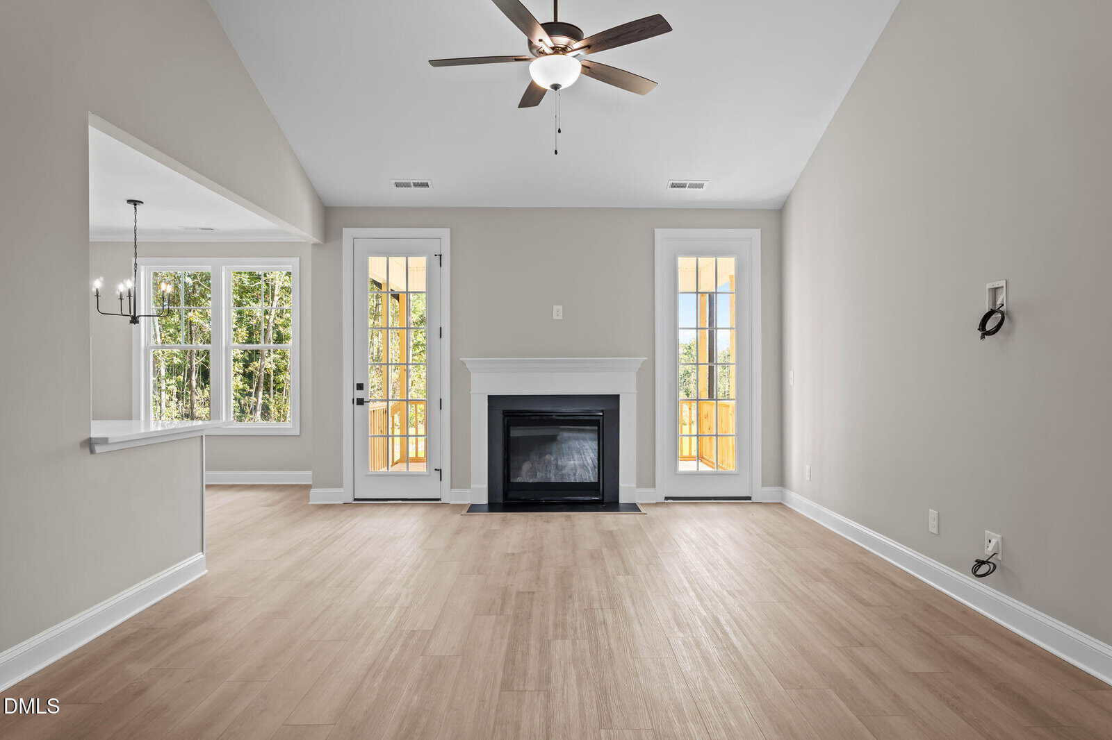 Bright living room with light wood floors, a ceiling fan, a fireplace centered between two glass doors, and an open view to the dining area and kitchen. Walls are painted light gray.