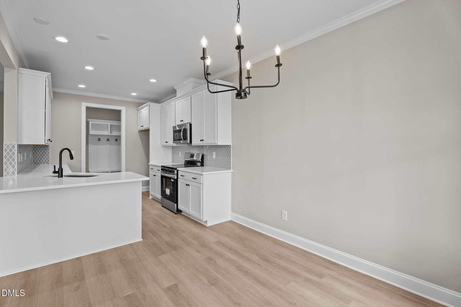Modern kitchen and dining area with white cabinets, stainless steel appliances, a center island with sink, light wood flooring, beige walls, and a black chandelier. Open layout with laundry area visible in the background.