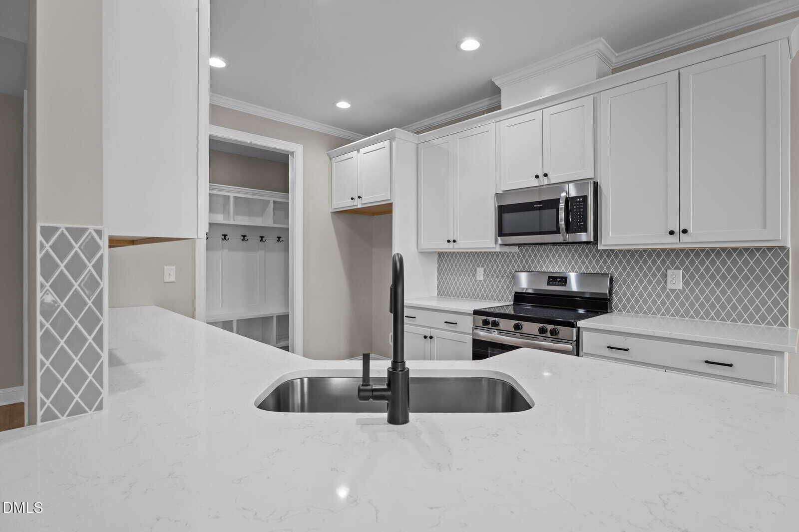 Modern kitchen with white cabinets, stainless steel appliances, a built-in microwave, patterned tile backsplash, and a large white island featuring a sink with a black faucet. A mudroom with hooks is visible in the background.