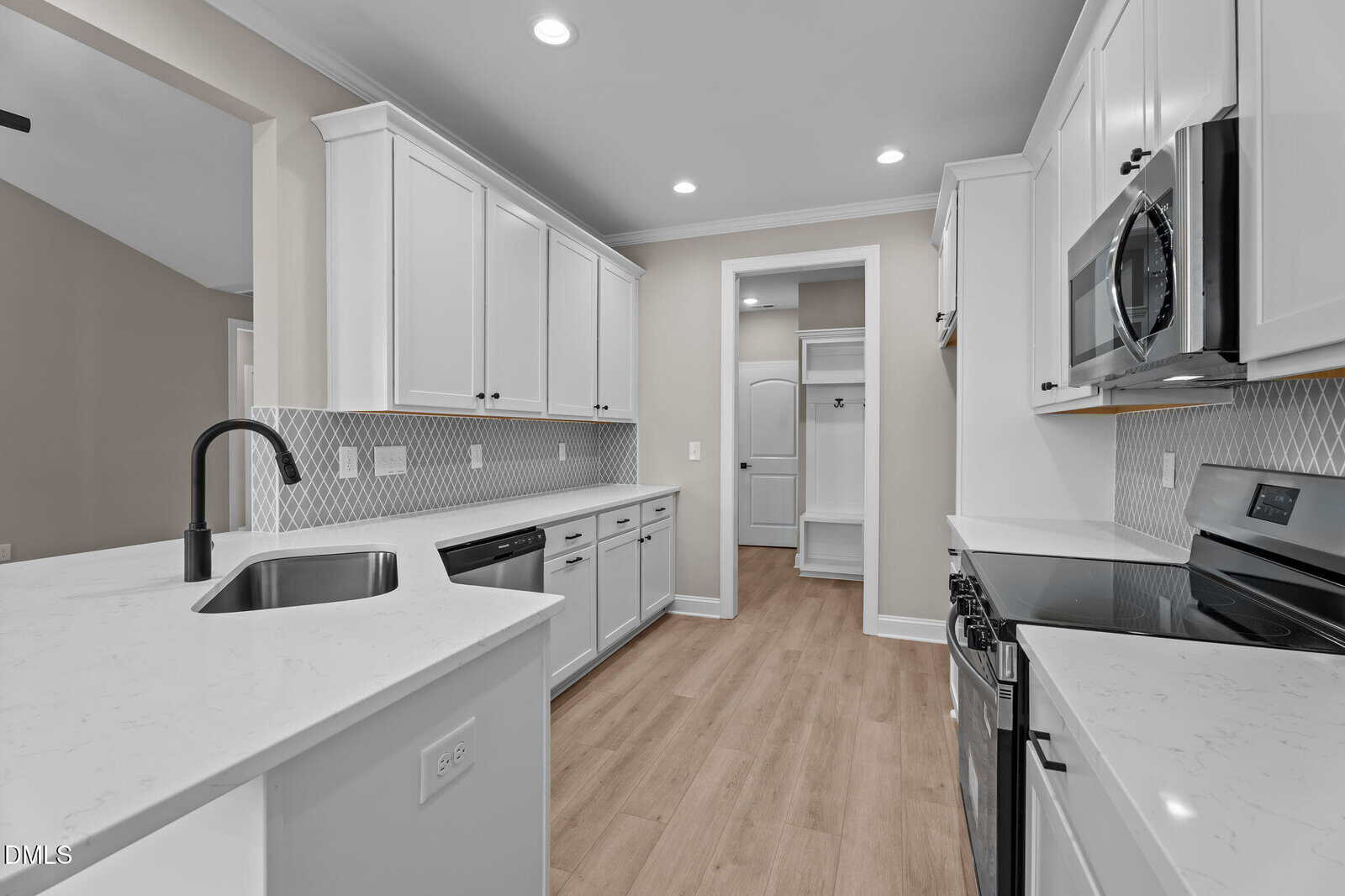 Modern kitchen with white cabinets, marble countertops, stainless steel appliances, black fixtures, light wood flooring, and geometric backsplash. A hallway with built-in storage is visible in the background.