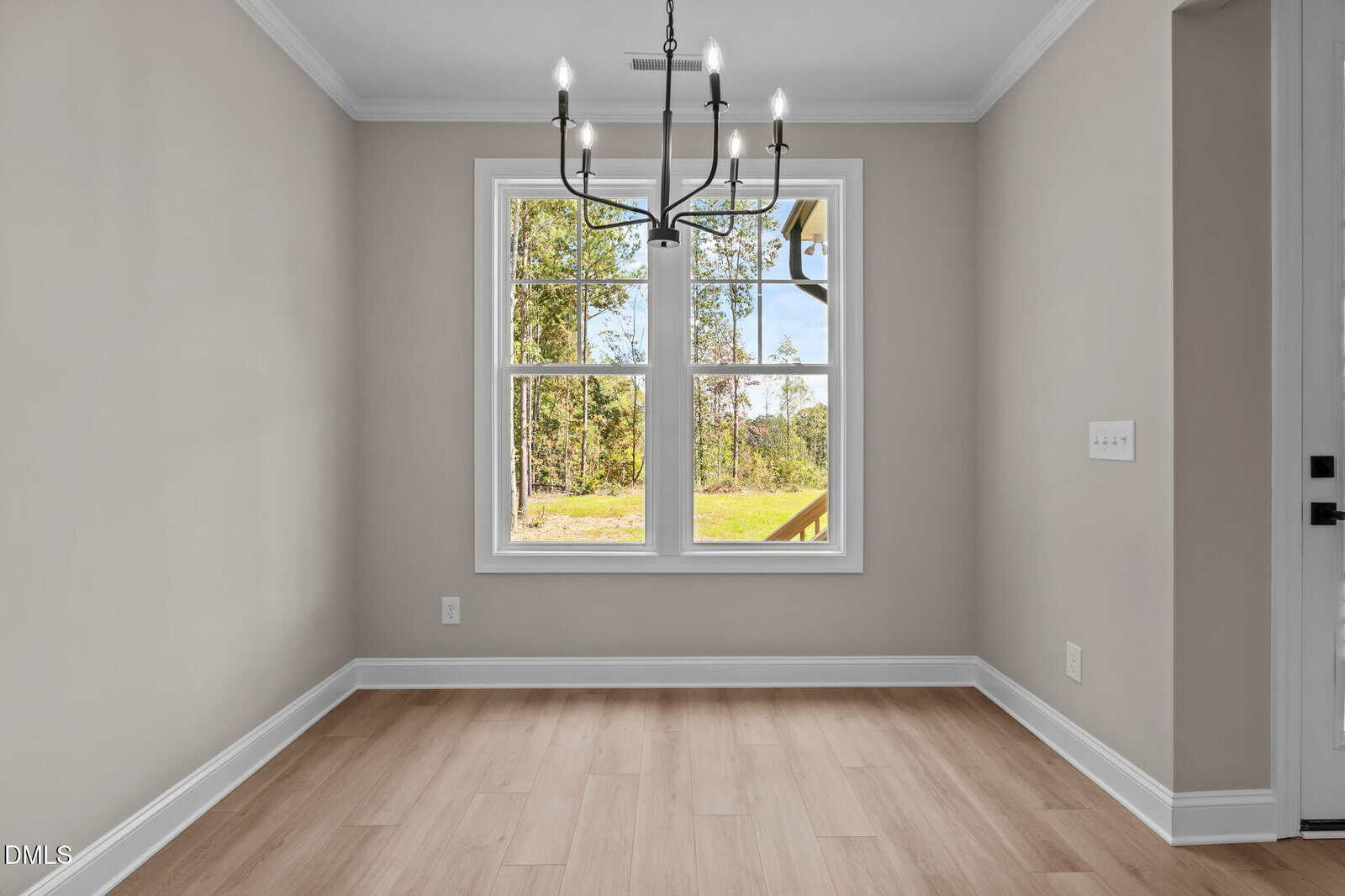 A small, empty dining area with light wood flooring, beige walls, a modern black chandelier, and a large window overlooking trees and a grassy yard. Natural light fills the room.